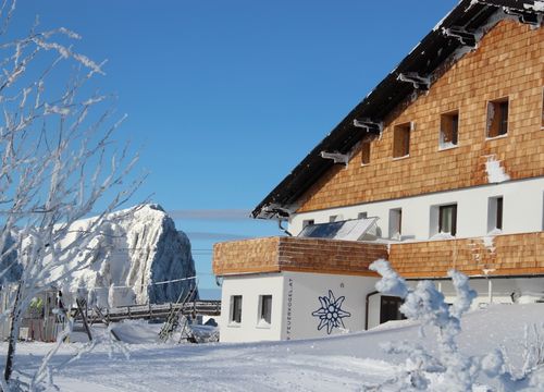 Snow-covered Berggasthof Edelweiss with wooden elements, surrounded by frosted trees and mountain peaks beneath a blue sky.