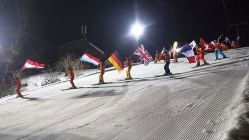 Skifahrer in farbenfrohen Kostümen fahren mit verschiedenen Nationalflaggen einen nächtlichen, von hellen Lichtern beleuchteten Schneehang hinunter.
