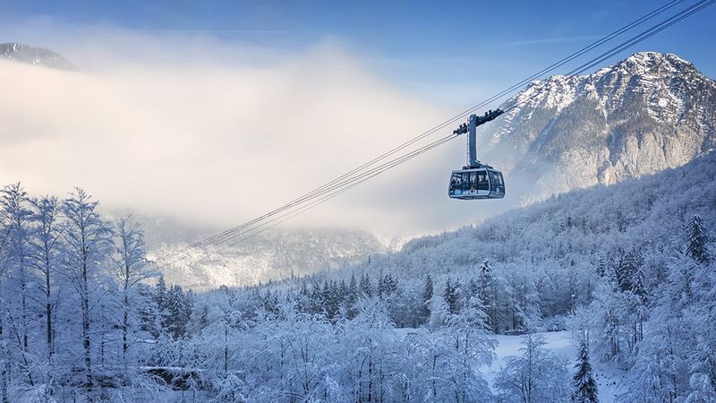 Seilbahn schwebt über verschneite Bäume und nebelverhangene Berge bei klarem Winterwetter am Dachstein Krippenstein.