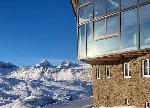 Moderne Berghütte mit Glasfassade, Sonnenaufgang spiegelt sich, dahinter schneebedeckte Gipfel und frisch präparierte Skipiste.