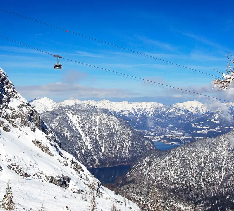 Eine Bergbahn überquert eine verschneite Berglandschaft mit Blick auf tief eingeschnittene Täler und ferne Gipfel.