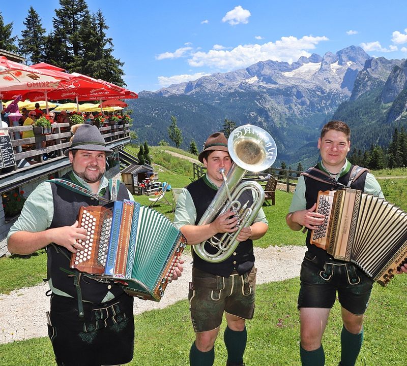 Drei Männer in traditioneller Kleidung spielen Akkordeons und eine Posaune im Freien, mit Dachstein Gletscher und einer Hütte im Hintergrund. Ein sonniger Tag.