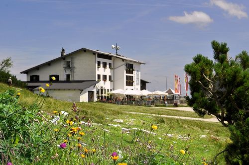 Weißes Feuerkogelhaus mit Außensitzplätzen und Sonnenschirmen auf einer Wiese mit Wildblumen unter strahlend blauem Himmel.