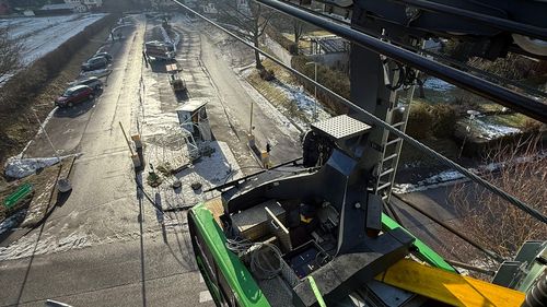 Blick aus der Seilbahn aus großer Höhe auf die grüne Kabine, die komplexe Seilbahnanlage, eine Straße darunter und einen sonnigen, schneebedeckten Berg.