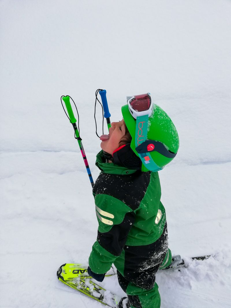 A child in green ski wear and a helmet, playfully catching snowflakes on their tongue and standing on the snow-covered ground with ski poles.