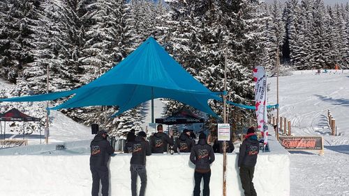 Menschen in dunkler Winterkleidung stehen an einer schneebedeckten Bar unter einem blauen Zelt, umgeben von verschneiten Bäumen und einem klaren Himmel.