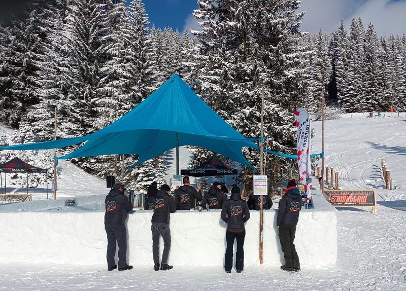Menschen in dunkler Winterkleidung stehen an einer schneebedeckten Bar unter einem blauen Zelt, umgeben von verschneiten Bäumen und einem klaren Himmel.