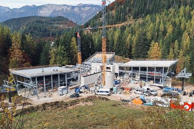 Baustelle einer Seilbahnstation in einem bergigen Waldgebiet mit Kränen, Fahrzeugen und herbstlichem Laub in der Umgebung.