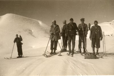 Sechs Skifahrer in nostalgischer Kleidung stehen auf verschneitem Hang, eine Person posiert scherzhaft – Szene vom Feuerkogel.