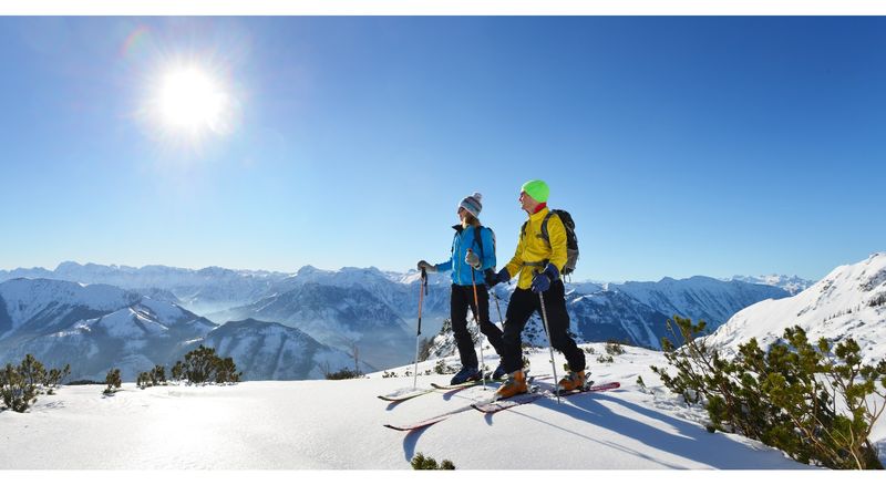 Zwei Menschen steigen auf Tourenski bei strahlendem Sonnenschein durch die verschneite Bergwelt und genießen die Aussicht.
