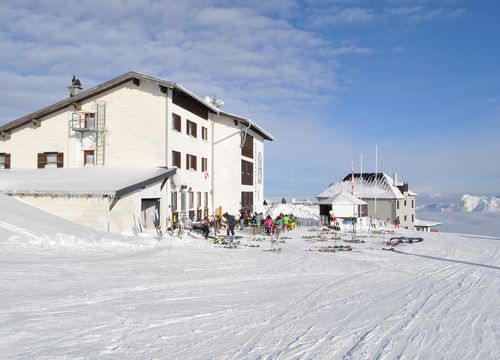 Snow-covered Feuerkogelhaus with skiers and equipment in front, surrounded by mountains under a clear blue sky.