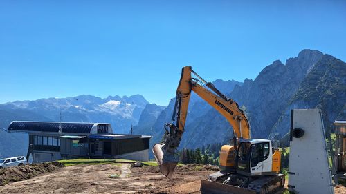 Excavator at work on a construction site with mountains in the background under a bright blue sky.