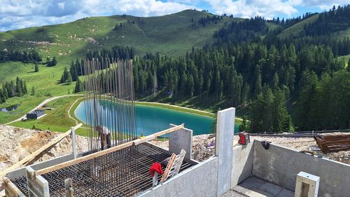 Baustelle mit Stahlbewehrung und Betonformen im Vordergrund, mit Blick auf eine grüne Landschaft mit Hügeln, Bäumen und einem kleinem Teich unter blauem Himmel.