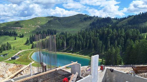 Construction site with steel reinforcement and concrete forms in the foreground, overlooking a green landscape with hills, trees and a small pond under a blue sky.