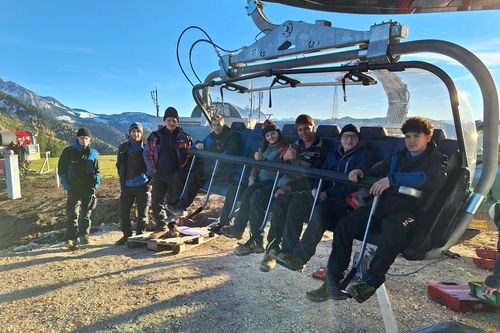 Gruppe von jungen Menschen, die in einer Bergbahnstation auf einem Sessellift sitzt, mit schneebedeckten Gipfeln, grünen Wiesen und strahlend blauem Himmel im Hintergrund.