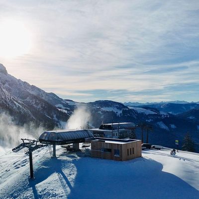 Verschneite Berglandschaft mit einer Skiliftstation, Kiefern und strahlendem Sonnenschein über fernen Gipfeln unter einem teilweise bewölkten Himmel.