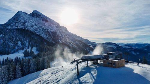 Verschneite Berglandschaft mit einer Skiliftstation, Kiefern und strahlendem Sonnenschein über fernen Gipfeln unter einem teilweise bewölkten Himmel.