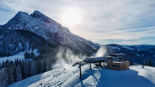Verschneite Berglandschaft mit einer Skiliftstation, Kiefern und strahlendem Sonnenschein über fernen Gipfeln unter einem teilweise bewölkten Himmel.