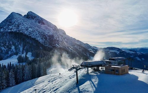 Verschneite Berglandschaft mit einer Skiliftstation, Kiefern und strahlendem Sonnenschein über fernen Gipfeln unter einem teilweise bewölkten Himmel.