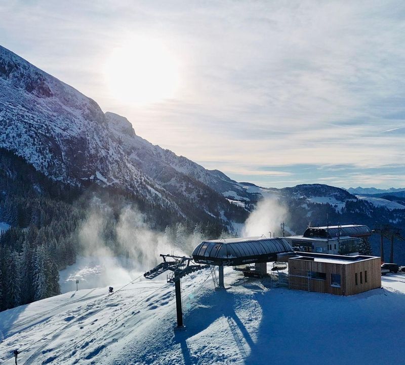 Snowy mountain landscape with a ski lift station, pine trees, and a bright sun shining over distant peaks under a partly cloudy sky.