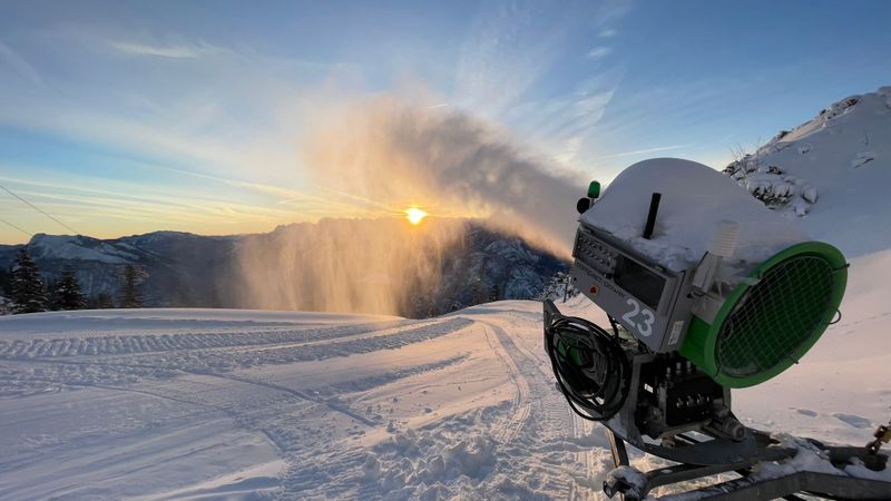 Schneekanone pustet feinen Schnee in die Luft, während die Sonne über verschneiten Bergen aufgeht.