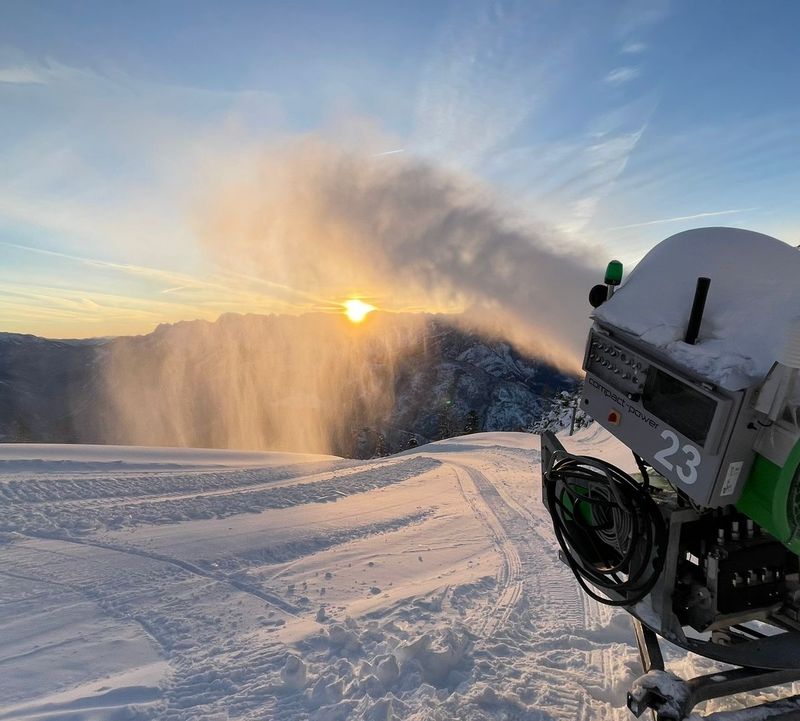 Schneekanone pustet feinen Schnee in die Luft, während die Sonne über verschneiten Bergen aufgeht.