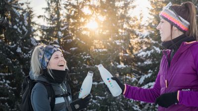 Zwei Personen in Winterkleidung stoßen mit Wasserflaschen an und lächeln vor verschneitem Wald und sonnigem Hintergrund.