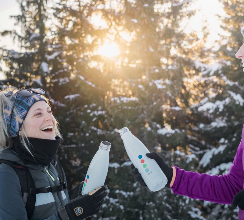 Zwei Personen in Winterkleidung stoßen mit Wasserflaschen an und lächeln vor verschneitem Wald und sonnigem Hintergrund.