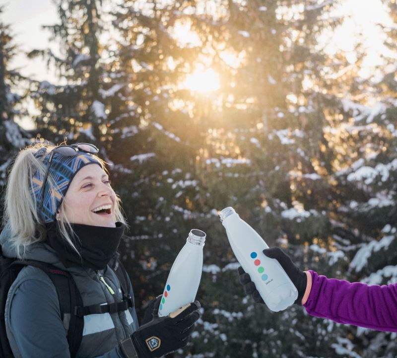 Zwei Personen in Winterkleidung stoßen mit Wasserflaschen an und lächeln vor verschneitem Wald und sonnigem Hintergrund.