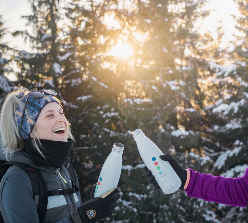 Two people in winter clothing clink water bottles and smile in front of a snowy forest and sunny background.