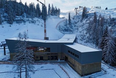 Snow-covered industrial building surrounded by pine trees and hills, with a crane and ski lift in the background under a cloudy sky.