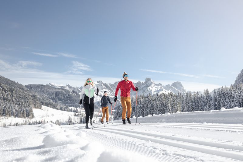 Drei Personen beim Skilanglauf auf einer verschneiten Loipe, umgeben von schneebedeckten Bäumen und Bergen unter einem klaren blauen Himmel.