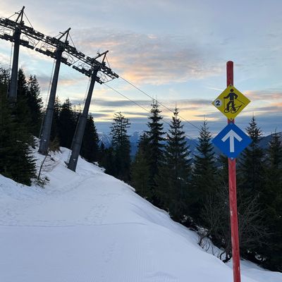 A snow-covered mountain trail with a ski lift on the left and a sign pointing to a snowshoe trail, surrounded by evergreen trees and a cloudy sky.