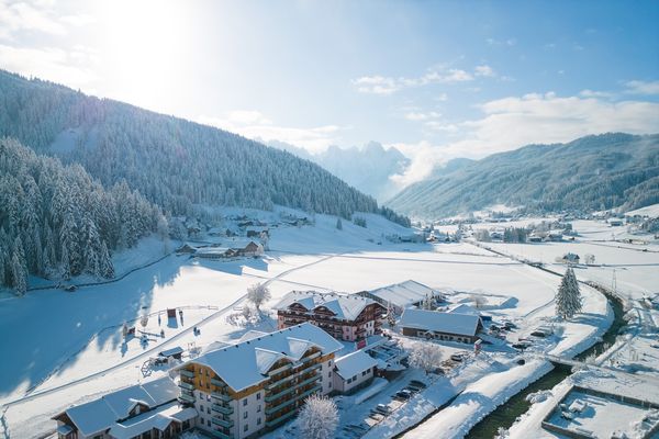 Luftaufnahme eines verschneiten Berghotels mit sonnenbeschienenen Hängen, bewaldeten Hügeln und einem kleinen Bach, der durch die Landschaft fließt.