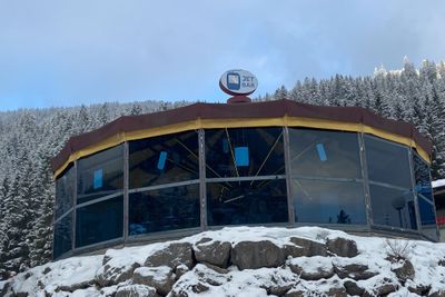 Ein rundes Gebäude mit großen Fenstern und der Aufschrift „Jet Bar“ steht auf einem verschneiten Hügel, umgeben von schneebedeckten Bäumen und unter einem bewölkten Himmel.