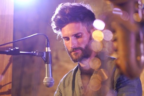 Ein bärtiger Musiker spielt konzentriert eine Handpan unter lila Bühnenlicht, ein Mikrofon steht davor. Rechts sanfte Bokeh-Effekte.