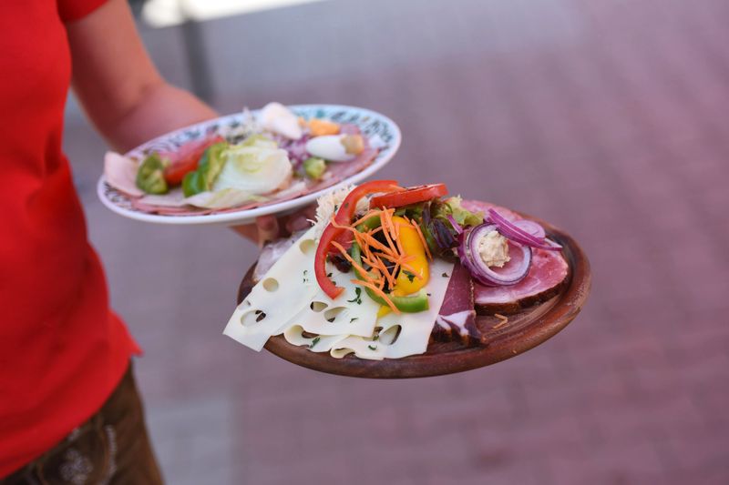 Person trägt zwei Teller mit Fleisch, Käse, Gemüse und Garnitur vor verschwommenem Hintergrund.