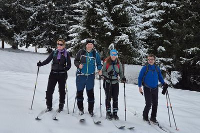 Four ski tourers are standing in front of a coniferous forest in a snow-covered landscape.