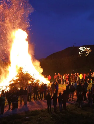 Menschen stehen um großes Sonnwendfeuer, darüber steigen Funken auf, am Hang leuchtet eine Figur aus Lichtern.