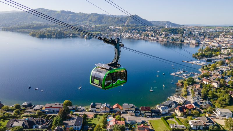 Die Grünberg Seilbahn fährt, im Hintergrund der Traunsee mit zahlreichen Segelbooten, umgeben von grünen Hügeln und der Stadt Gmunden.