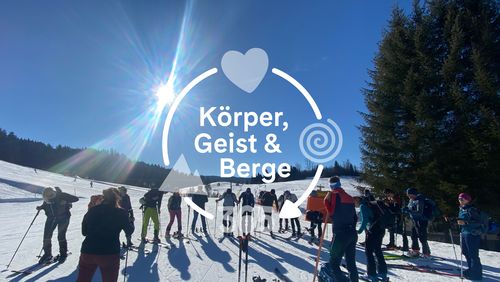 A group of people stand at the edge of a sunny, snow-covered ski slope surrounded by trees. The text ‘Body, Mind & Mountains’ is overlaid with a heart and spiral symbol.