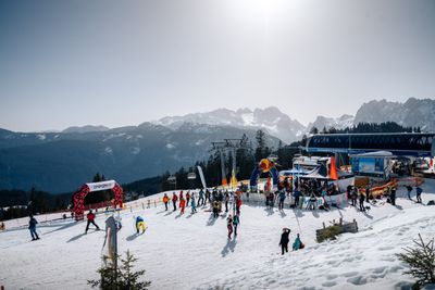 Skifahrer und Snowboarder bei der Bergstation eines Sessellifts mit Blick auf die umliegenden Berge, Veranstaltungszelten und einem roten Torbogen.