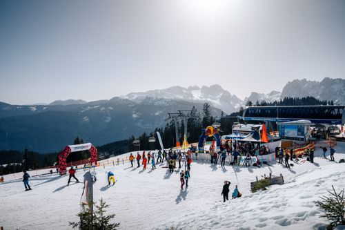 Skifahrer und Snowboarder bei der Bergstation eines Sessellifts mit Blick auf die umliegenden Berge, Veranstaltungszelten und einem roten Torbogen.