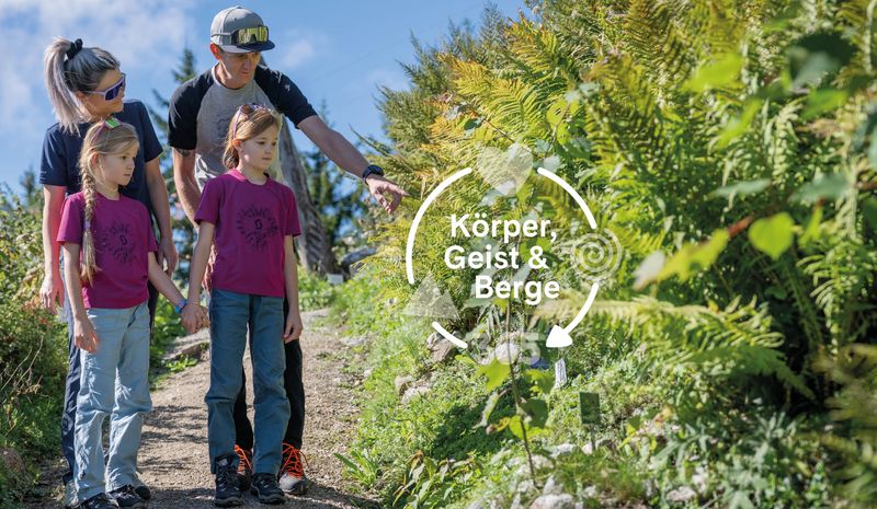 Eine Familie spaziert auf einem sonnigen Weg, wobei ein Mann auf Pflanzen zeigt. Bildunterschrift: „Körper, Geist & Berge“.