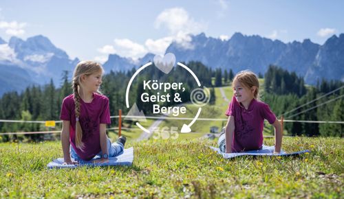 Two girls stretch on mats in a sunny field with mountains. Overlay: "Körper, Geist & Berge" (Body, Mind & Mountains) & "365".