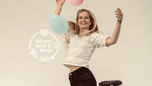 A smiling blonde woman is skipping around happily, holding pink and blue balloons in her hands. The text reads: “Body, Mind & Mountains 365”.