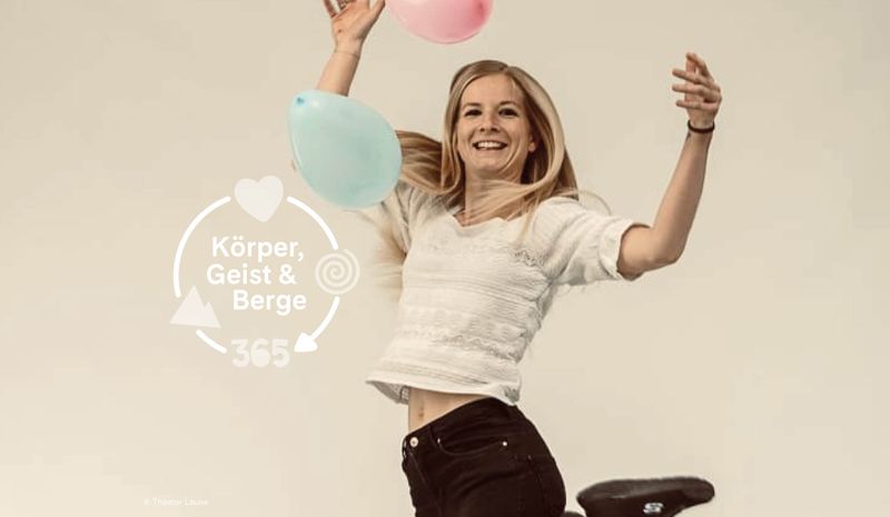 Eine lächelnde blonde Frau hüpft fröhlich herum und hält rosa und blaue Luftballons in den Händen. Der Text lautet: „Body, Mind & Mountains 365“.