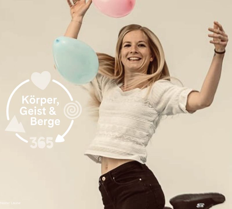 Eine lächelnde blonde Frau hüpft fröhlich herum und hält rosa und blaue Luftballons in den Händen. Der Text lautet: „Body, Mind & Mountains 365“.