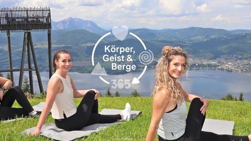 Two women are sitting in a twisted pose on mats on a grassy hill, smiling at the camera. A picturesque backdrop featuring a lake and mountains. Text: “Body, Mind & Mountains 365”.