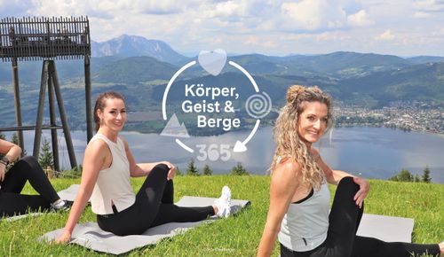 Zwei Frauen sitzen in einer Drehhaltung auf Matten auf einem grasbewachsenen Hügel und lächeln in die Kamera. Malerische Kulisse mit See und Bergen. Text: „Körper, Geist & Berge 365“.